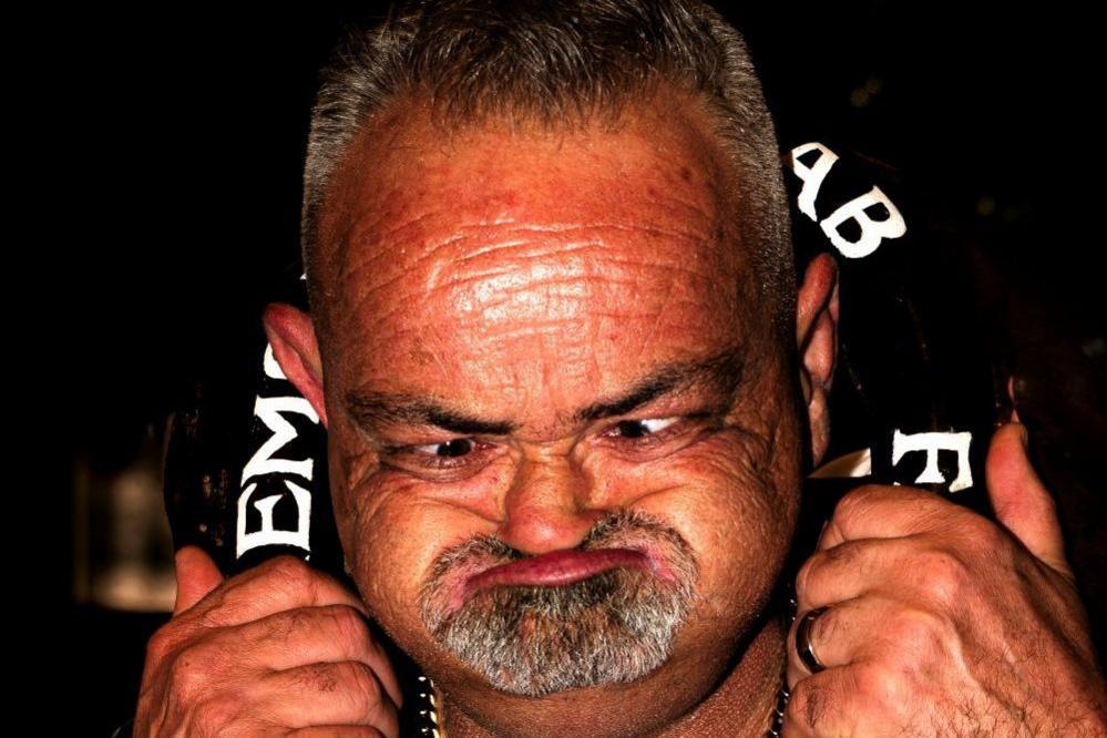 Close up of Ged Eccles distorting his face. His mouth is closed and his eyes look small and squashed. He has a grey goatee and has his hands placed on a large black horseshoe put around his neck. He is wearing a silver chain.