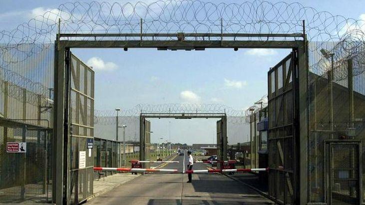 A prison guard in a white shirt and black trousers stands at a security gate in the inner area of the main entrance to Maghaberry prison where the main gates are open