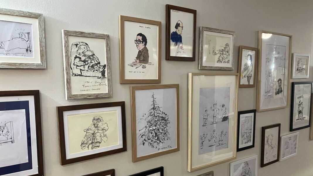 Artist, 94, thrilled by House of Commons exhibition A wall decorated in a care home with a large collection of framed artwork, primarily black-and-white cartoon sketches, caricatures, and ink drawings. The frames vary in size, color, and material, including light wood, dark wood, black, white, and weathered gray finishes.
