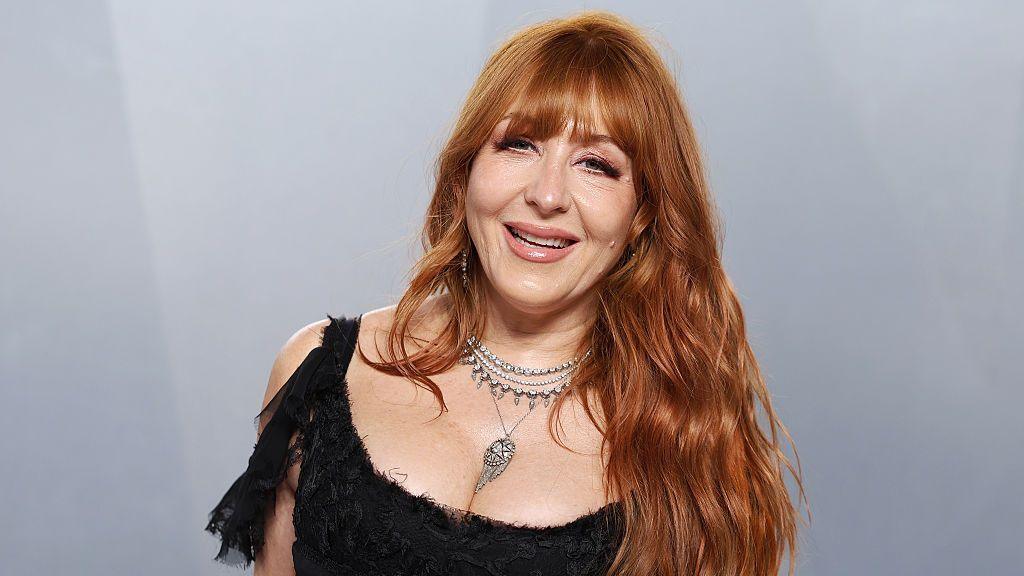 Charlotte Tilbury with long red hair, smiling, wearing a black textured top and silver necklaces