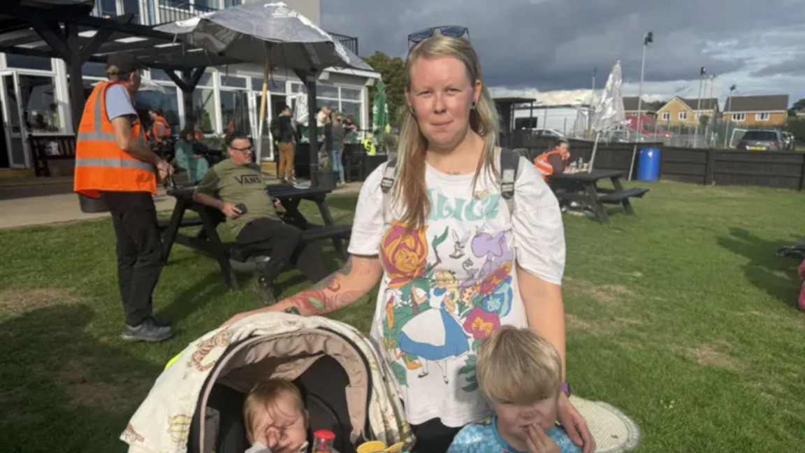 A woman with long blonde hair and sunglasses on her head standing behind her two children. One is in a pushchair and the other one is standing up. There are people sitting on a bench behind her and another man wearing a orange hi-vis jacket