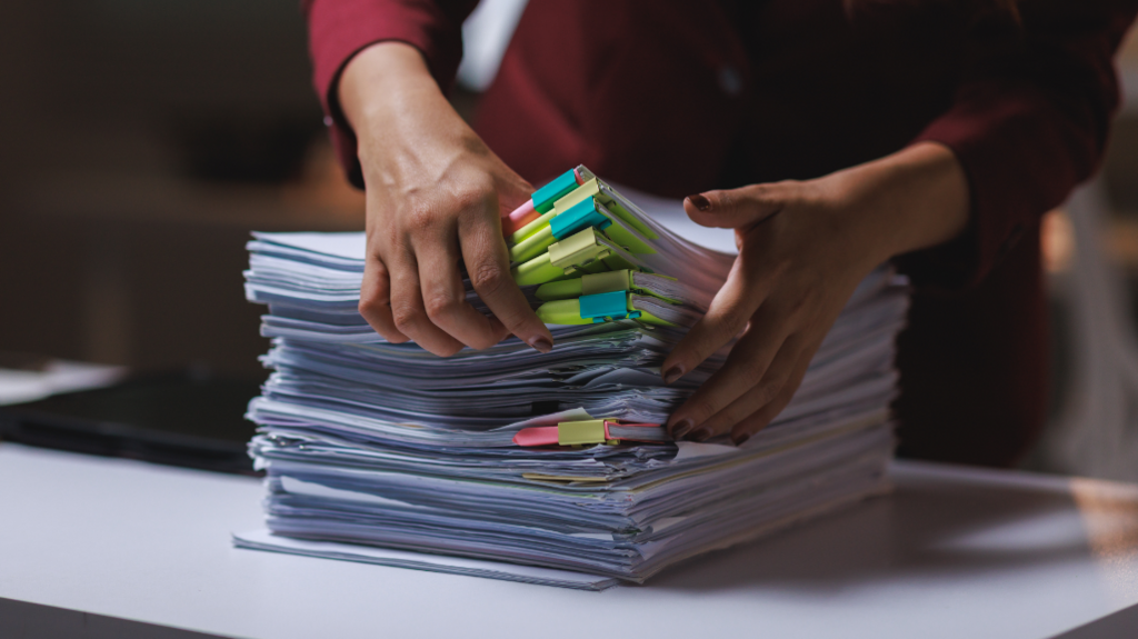 A pile of documents is stacked on a desk and a woman's hands are seen sorting through them.