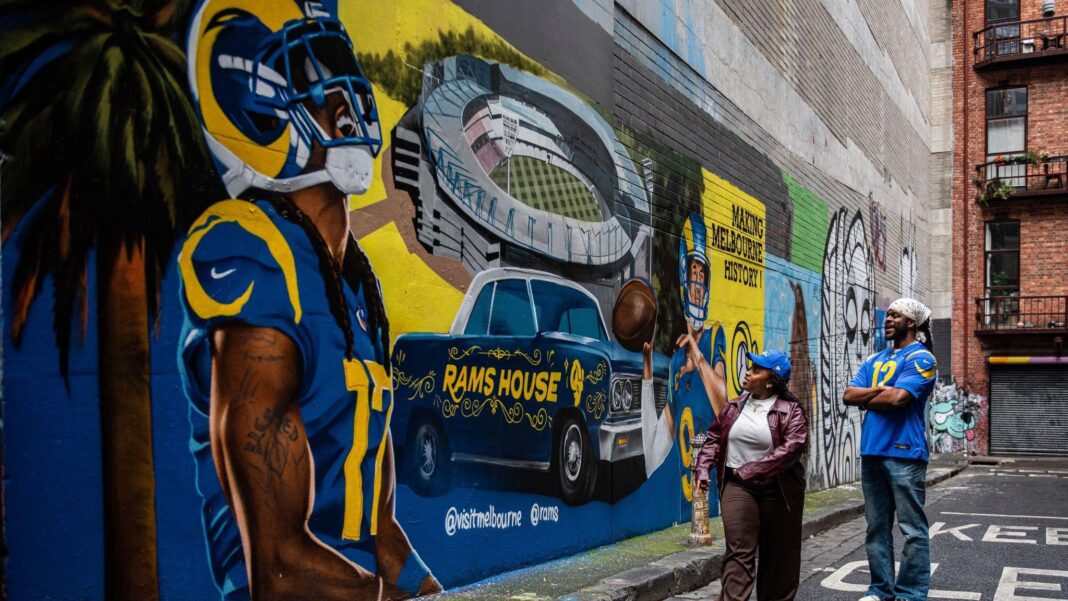 Australians love sport – but can the NFL win them over in debut Melbourne match? Two people in blue LA Rams gear admire a mural of the Rams featuring a player, a car and a stadium on the wall of a laneway