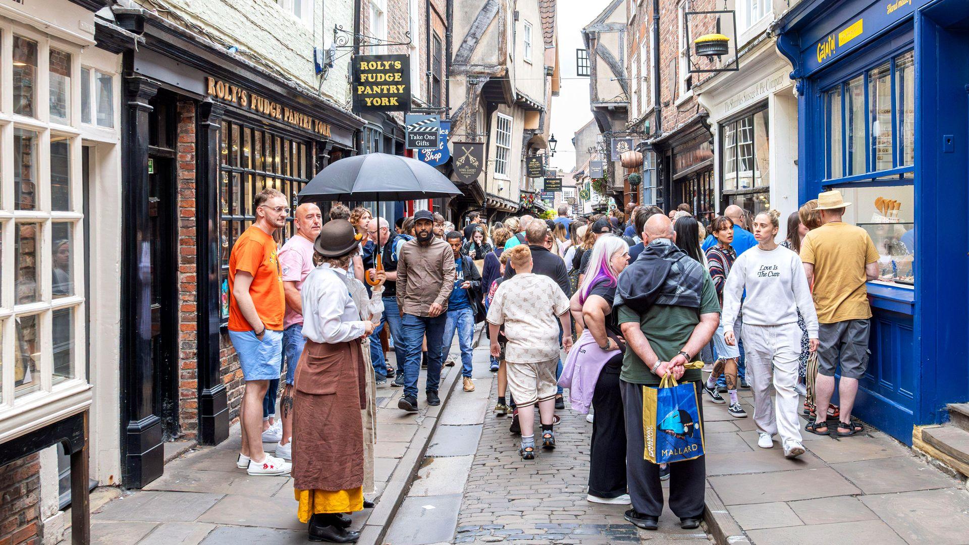 View along The Shambles in York, a narrow medieval street with timber-framed buildings housing shops and cafés. The scene shows the busy pedestrian area that retains its characteristic overhanging upper stories and historic layout.