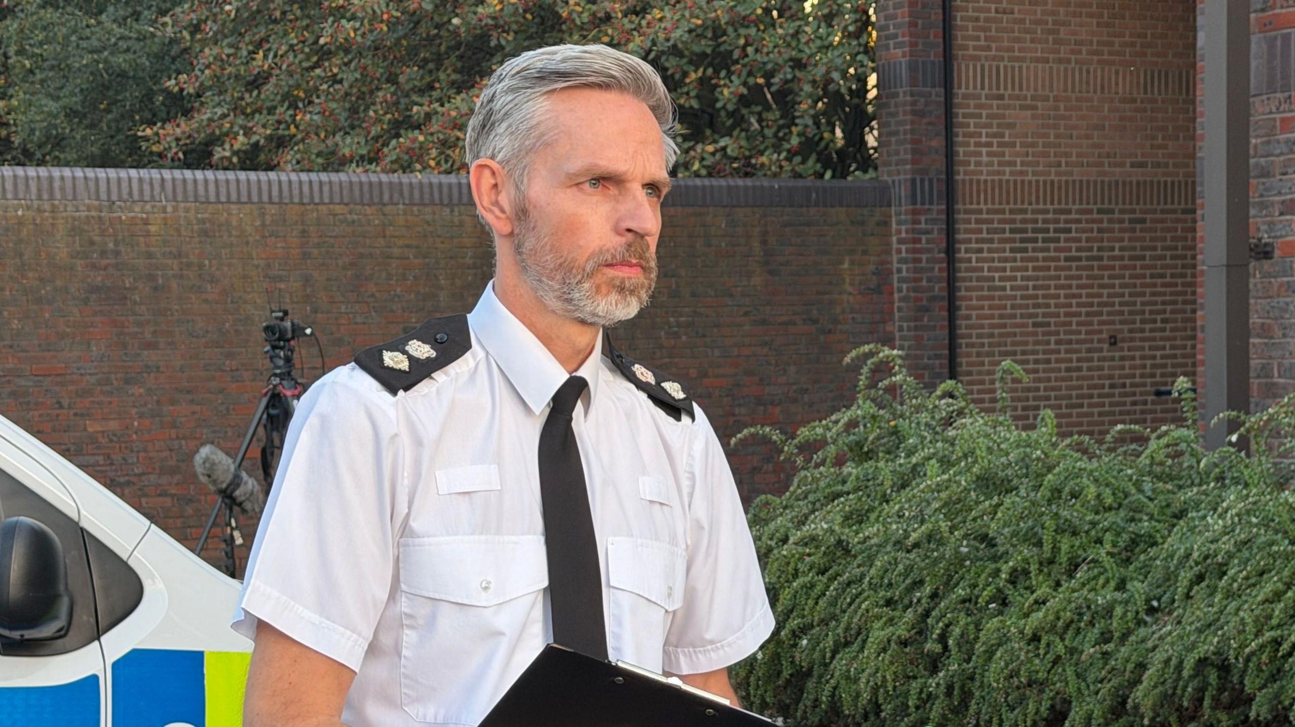 Scott Cowie looking to the right of the camera with a serious look on his face. He has short, grey hair, which has been styled to the side, and  a grey beard. He is wearing a white short-sleeved shirt with black epaulettes on his shoulders and a black tie. He is holding a black clipboard. Behind him a white, blue and yellow police van can just be seen as well as a large black camera stand and fluffy microphone. There is a green bush on the right.