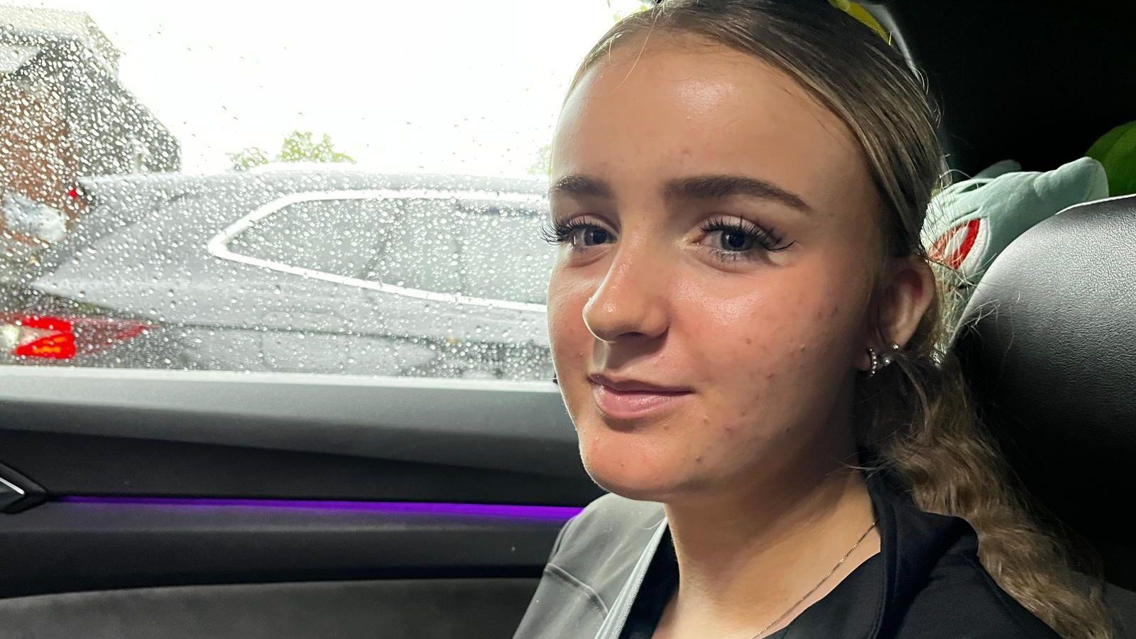 Lucy, a 19 year old woman, sitting in a car. There is rain on the window and a Pokemon teddy behind her