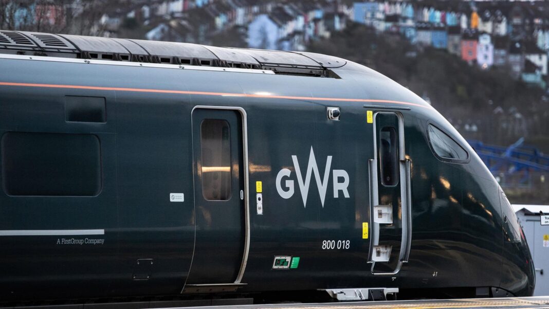 A green train at a platform, with colourful houses slightly out of focus in the background.