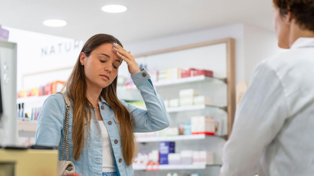 Migraines and acne among five more conditions pharmacists can treat Young woman holds her head as if she has a bad headache while standing in a pharmacy in front of a pharmacist wearing a white coat