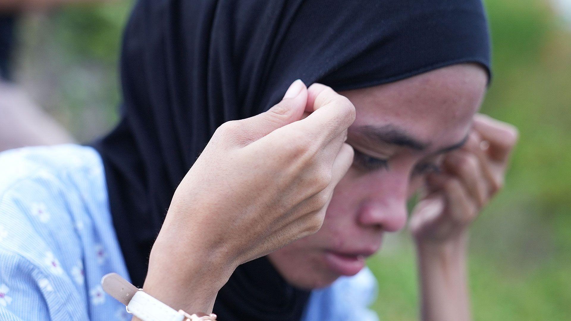 Sukma is hunched over as she adjusts her black hijab - she looks distressed. She is wearing a blue top with small white flowers.