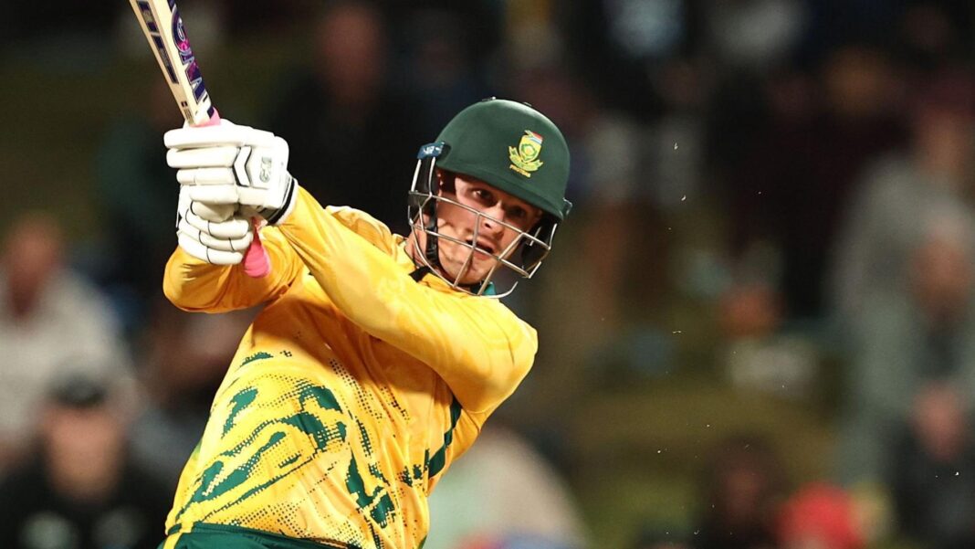 With a blurred crowd in the background, Jordan Hermann, in a yellow and green jersey and dark-coloured helmet, bats for South Africa, holding the bat up to his right with both hands