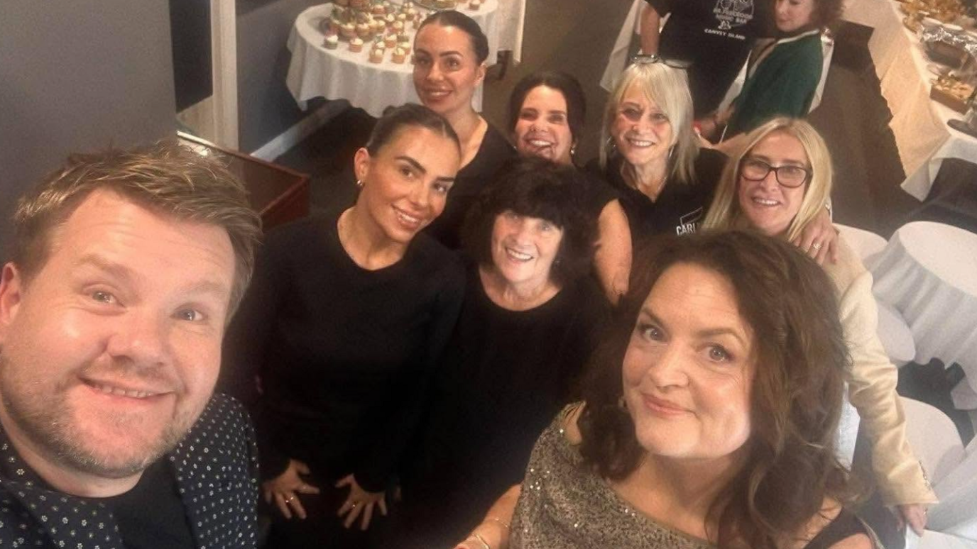 James Corden and Ruth Jones smiling towards the camera with staff members behind them. They are all smiling and wearing black shirts.