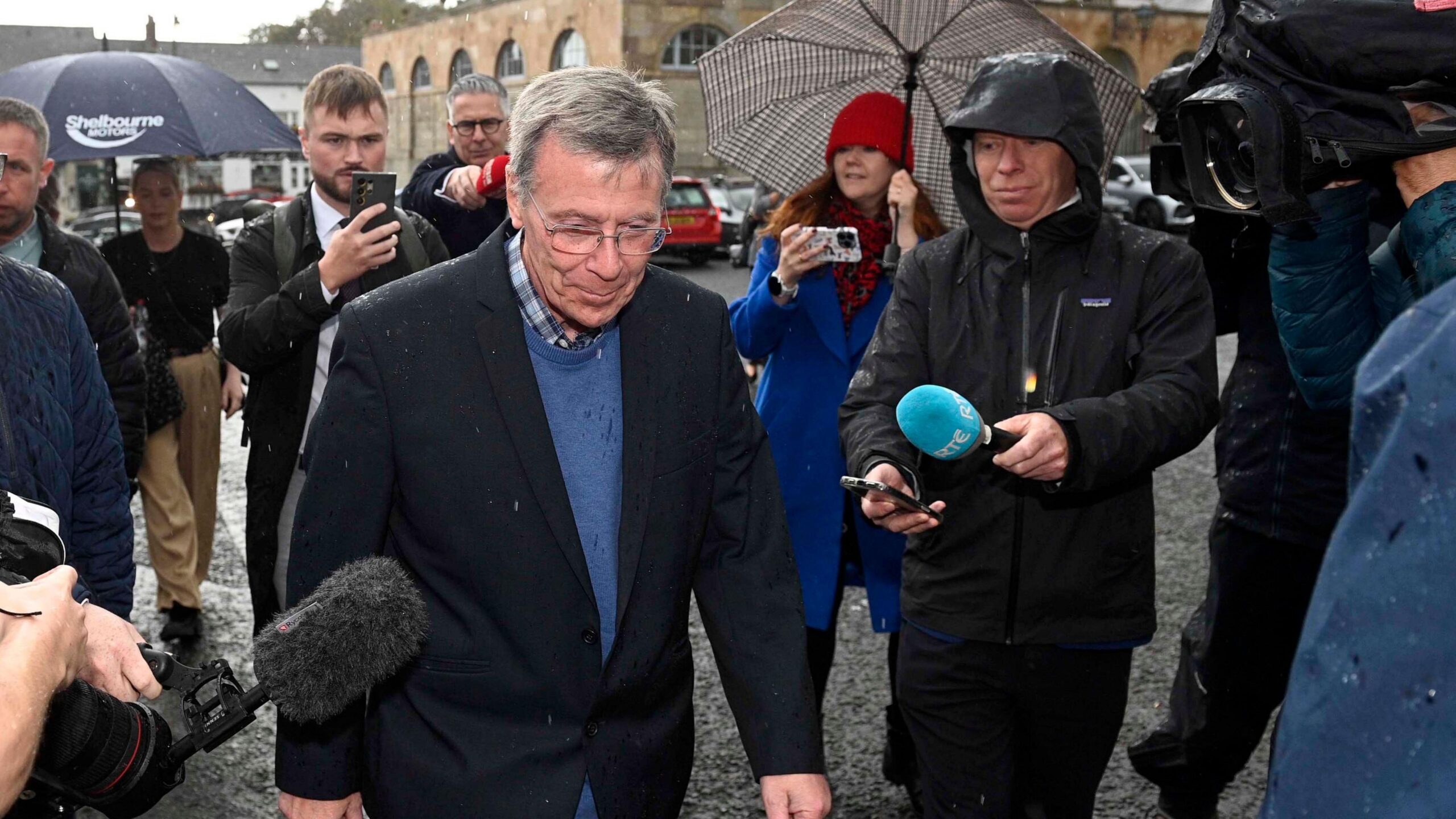 People are walking in the rain. Mics and cameras are pointing towards them. It's raining.