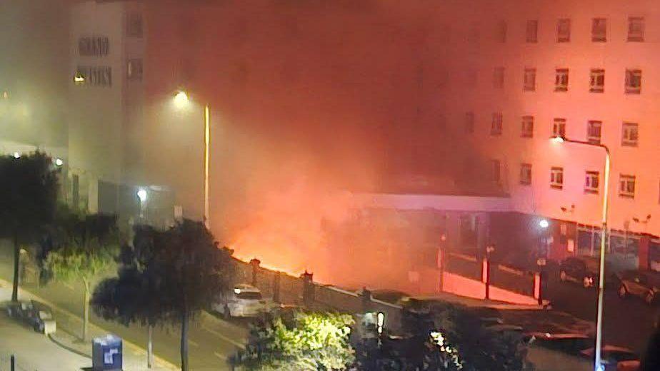 A fire in the basement of a hotel. Flames can be seen from below the building, and emergency services can be seen in the foreground.