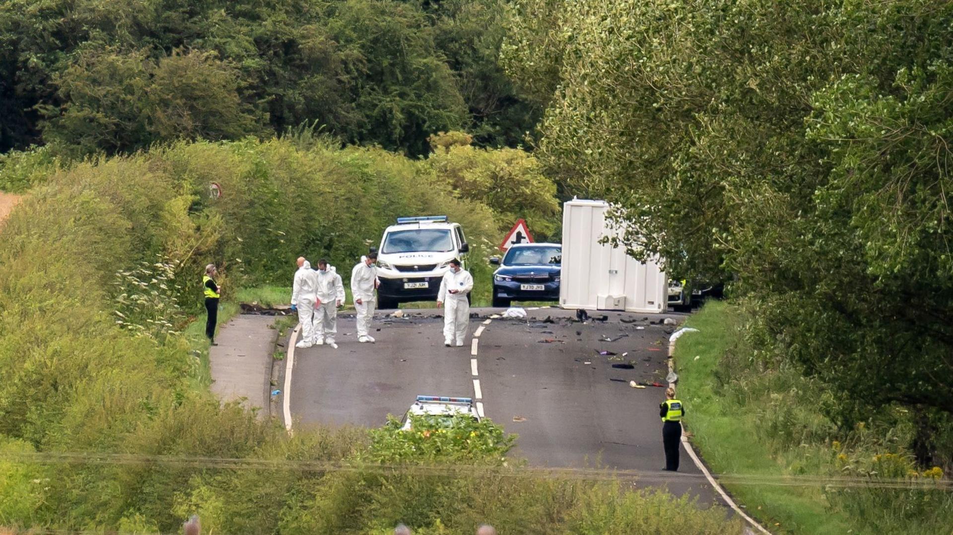 A long-distance view shows a rural road closed off while several people in white forensic suits examine an area containing scattered debris. Police vehicles and temporary structures are positioned on the road, with officers standing at different points around the scene. The road is bordered by fields, hedgerows and dense trees, with rolling countryside visible in the background.