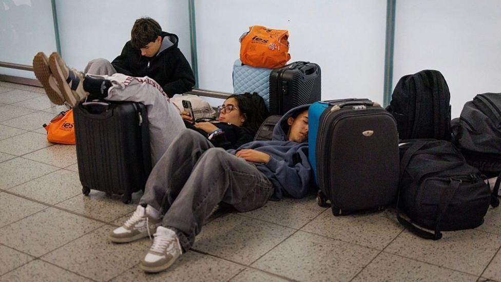 People laying on the floor in an airport with bags around them.