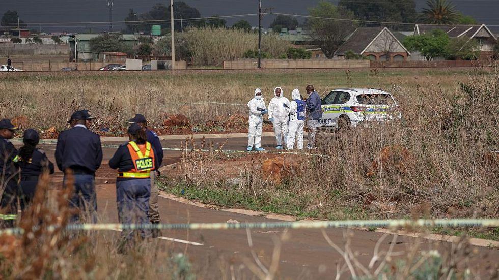 Members of the South African Police Service Forensic Pathology Services dressed in white protective gears work on a crime scene with police with their car nearby