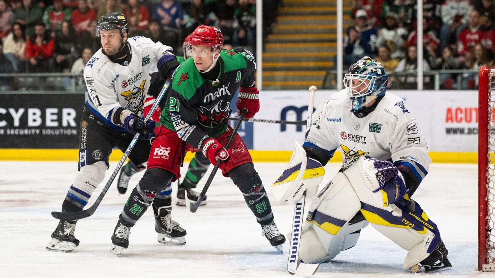 A Cardiff player has a Manchester Storm player to either side of him on the indoor rink, with a goal on the right hand side and spectators seen blurred out behind them.