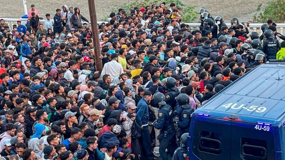 Hundreds of migrants crowd a narrow route in Ceuta as riot police form a cordon beside a police van during a relocation operation.