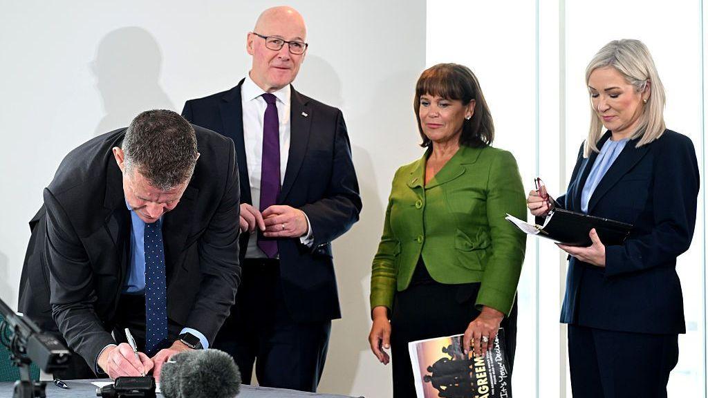 Four people, Michelle O'Neill, Mary Lou McDonald, Rhun ap Iorwerth and John Swinney, stand at a press conference, with ap Iorwerth bending down to sign a document