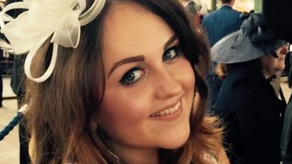 Charlotte Brown is seen smiling at the camera, wearing a white fascinator. Other  wedding guests are pictured in the background.