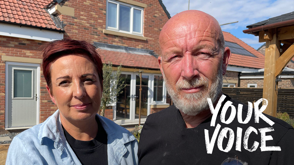 Elissa and Johnnie stand in front of a modern brick house with red‑tiled roofing and solar panels. Elissa wears a light blue shirt, the Johnnie a black shirt with safety glasses tucked into the pocket. The house has white‑framed windows, a light‑coloured door, and a clear blue sky overhead.