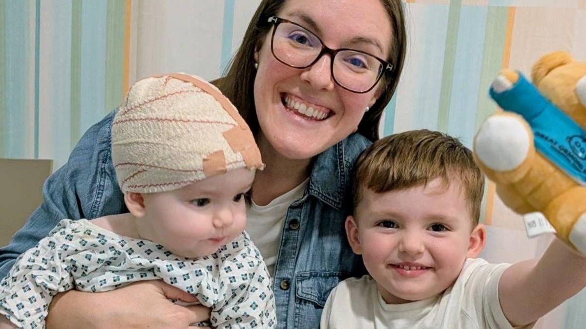 A woman wearing glasses sat smiling holding a baby with a bandage around his head and with her arm around a toddler who is holding a teddy.