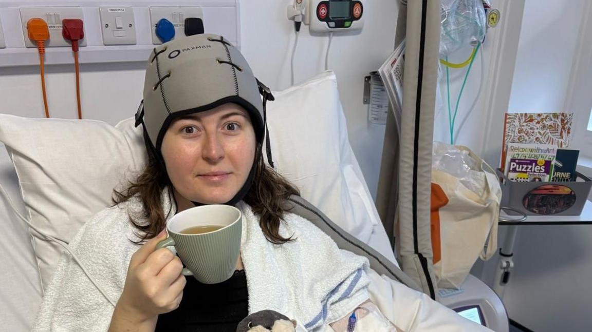 Fionnuala Quinlan-Bennett is seen in a hospital treatment area. She is sitting upright in a hospital bed. A cooling cap is fitted to her head and tubing is attached to medical equipment. A large mug, books and personal belongings are visible nearby. Medical monitoring equipment and hospital furnishings can be seen in the background.