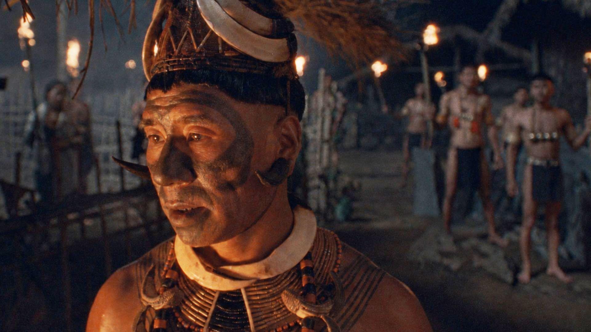 A man in traditional Konyak attire and facial tattoos stands in a village at night, illuminated by warm torchlight, with other figures visible in the background.