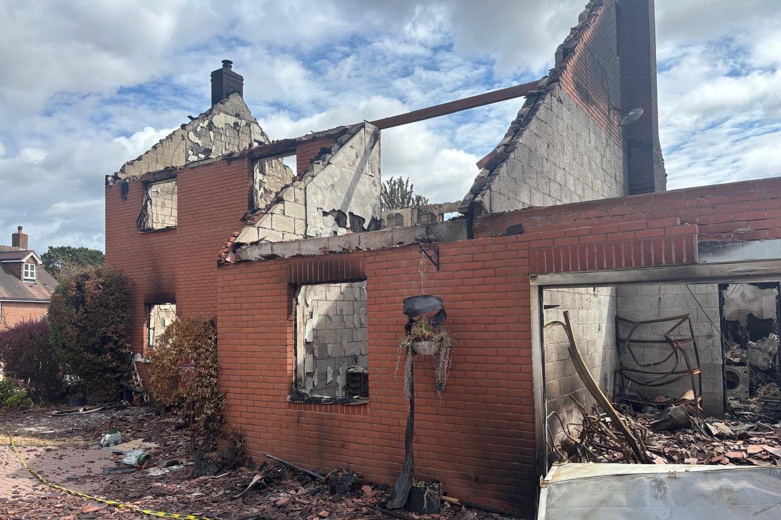 The ruins of a burnt out house. The roof is entirely gone while red brick walls remain with detritus all around.