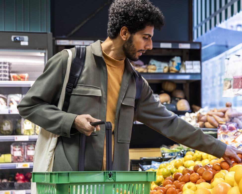 Man in a yellow t-shirt and grey jacket holding green shopping basket choosing lemon from crate in grocery store
