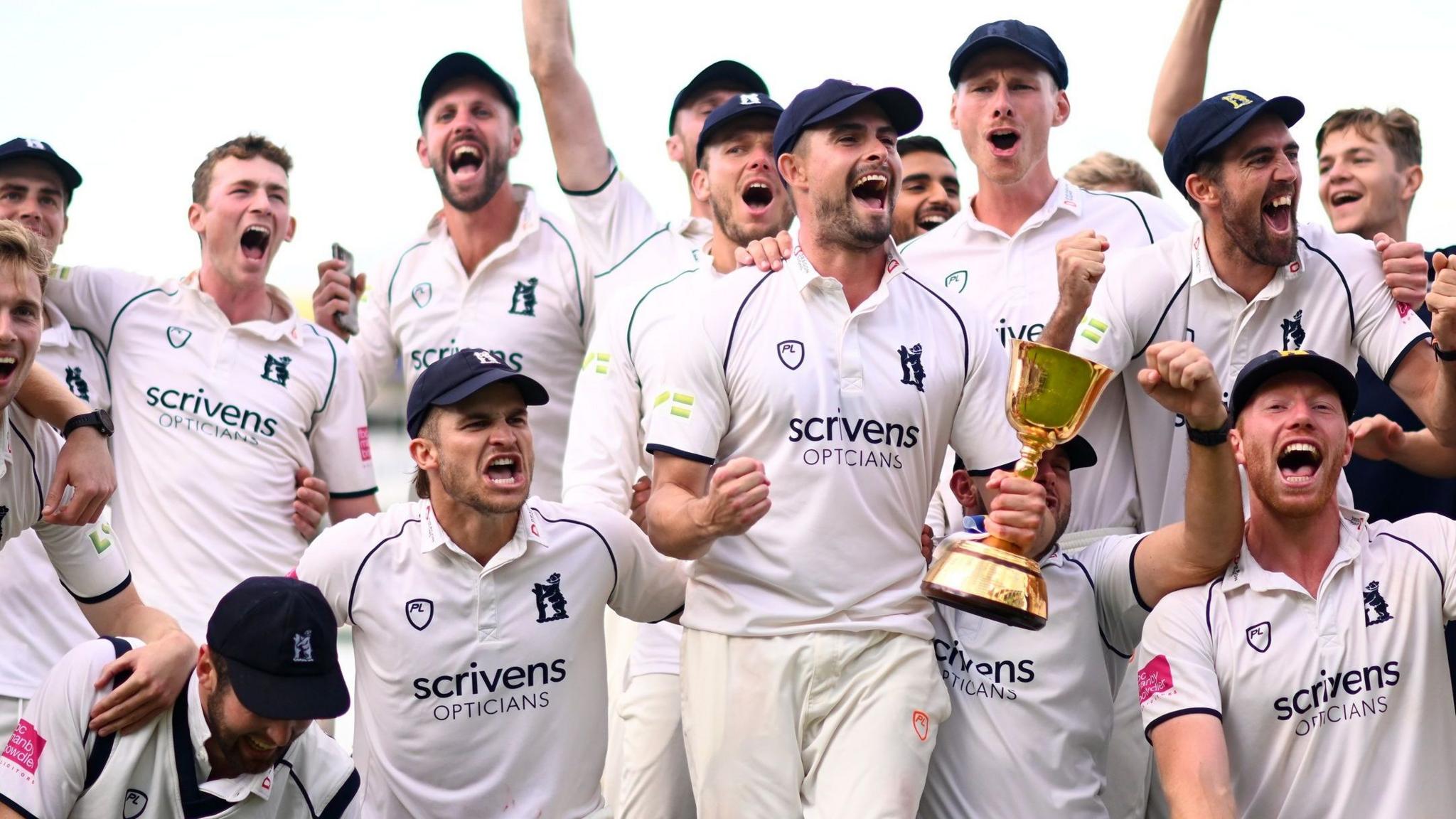Bears skipper Will Rhodes (centre) holds the trophy in his left hand and clenches his right fist, he is surrounded by team-mates in a crowd who are celebrating