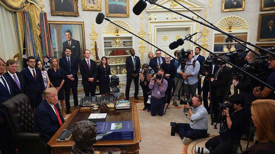 Trump seated at desk, flanked by officials and facing the press