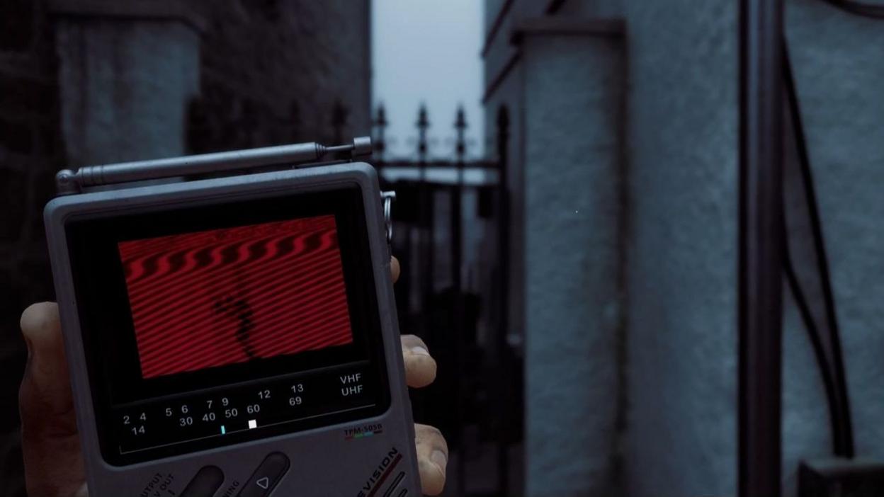 An in-game screenshot showing the handheld grey CRT monitor with a fuzzy red screen and a silhouette of a monster on it. The player is holding it, behind the view is a gate.