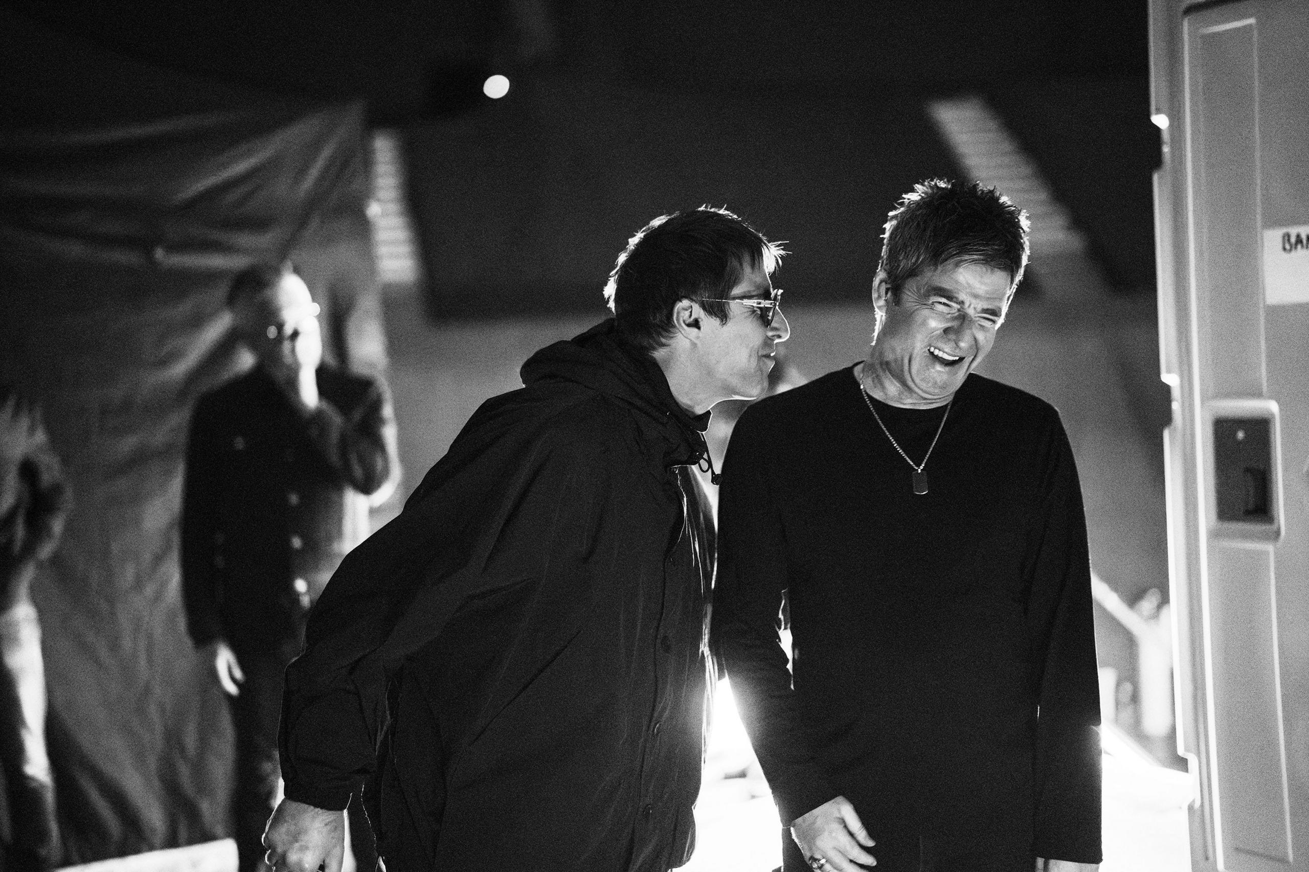 Liam and Noel Gallagher backstage during their 2025 reunion tour. Liam has on a trademark black cagoule and is saying something to Noel, who is laughing.