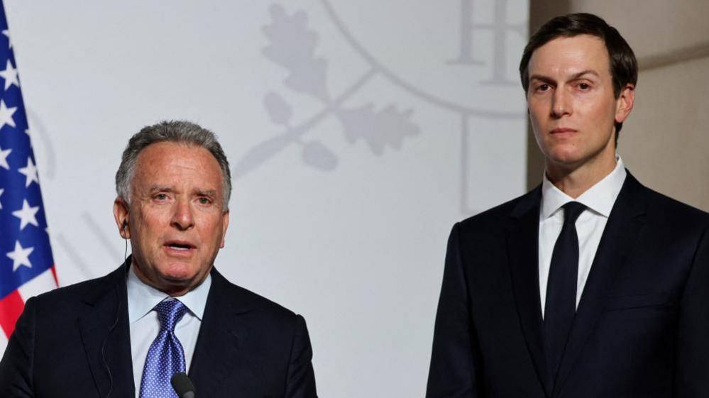 Steve Witkoff (left) and Jared Kushner (right) stand side by side looking directly towards the camera. They are both in black suits with white shirts. Witkoff has a blue patterned tie and Kushner has a black tie