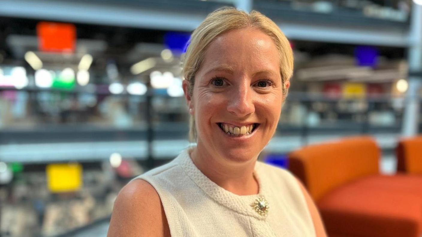 Michelle is smiling at the camera, she is wearing a cream sleeveless top and is sat on orange seats, with an open-plan, multi-level office setting in the background.