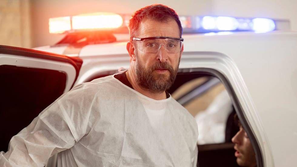 Noah Wyle in PPE glasses and shirt, in front of an ambulance with blue and orange lights flashing, in a scene from The Pitt
