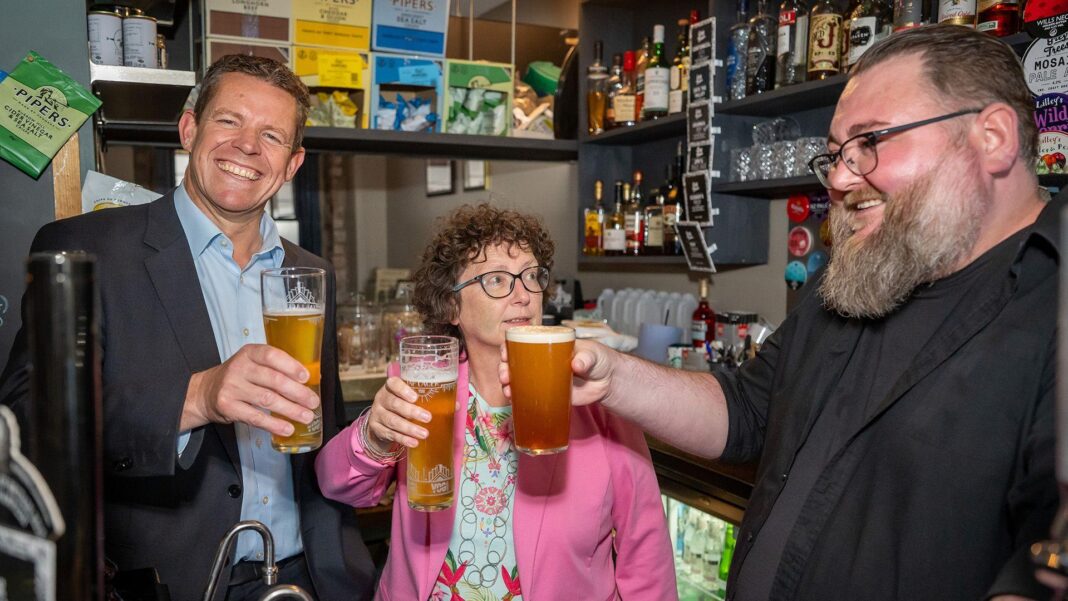 Rhun ap Iorwerth holding a pint and smiling at the camera. He has a dark grey suit and light blue shirt. Next to him is a woman with curly, brown hair and a pink blazer, with a light blue floral top underneath, she too holds a pint. Next to her is a man who smiles with a beard and glasses, and a black shirt, who also holds a pint. The three are stood behind a bar.
