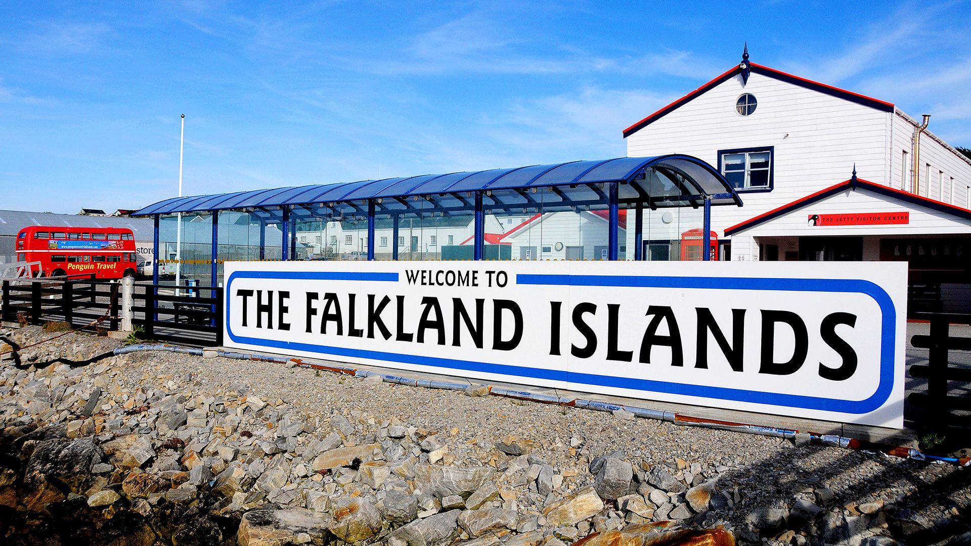 A sign saying 'welcome to The Falkland Islands' in front of a bus terminal at the harbour at Port Stanley in the Falklands Islands, with a red London-style bus in the background against a clear blue sky.