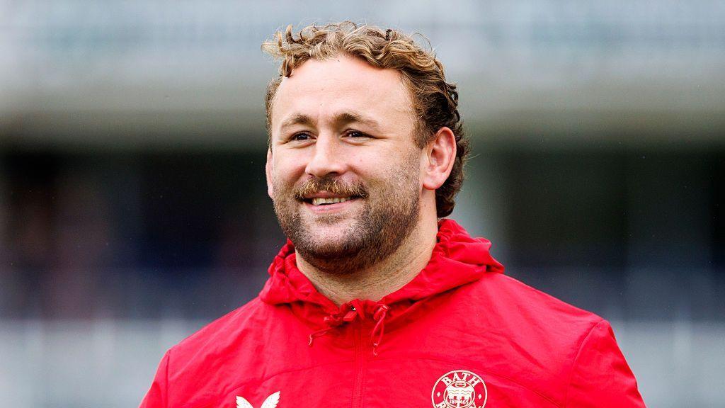 Will Stuart stands looking straight ahead smiling, wearing a red jacket with a Bath logo