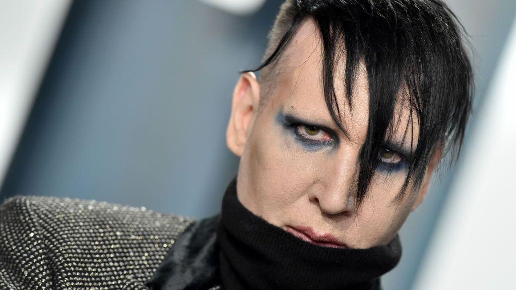 Marilyn Manson, real name Brian Warner, has black hair which covers some of his face. He has blue and black eye make on and wears a sparkly jacket with a black turtle neck.