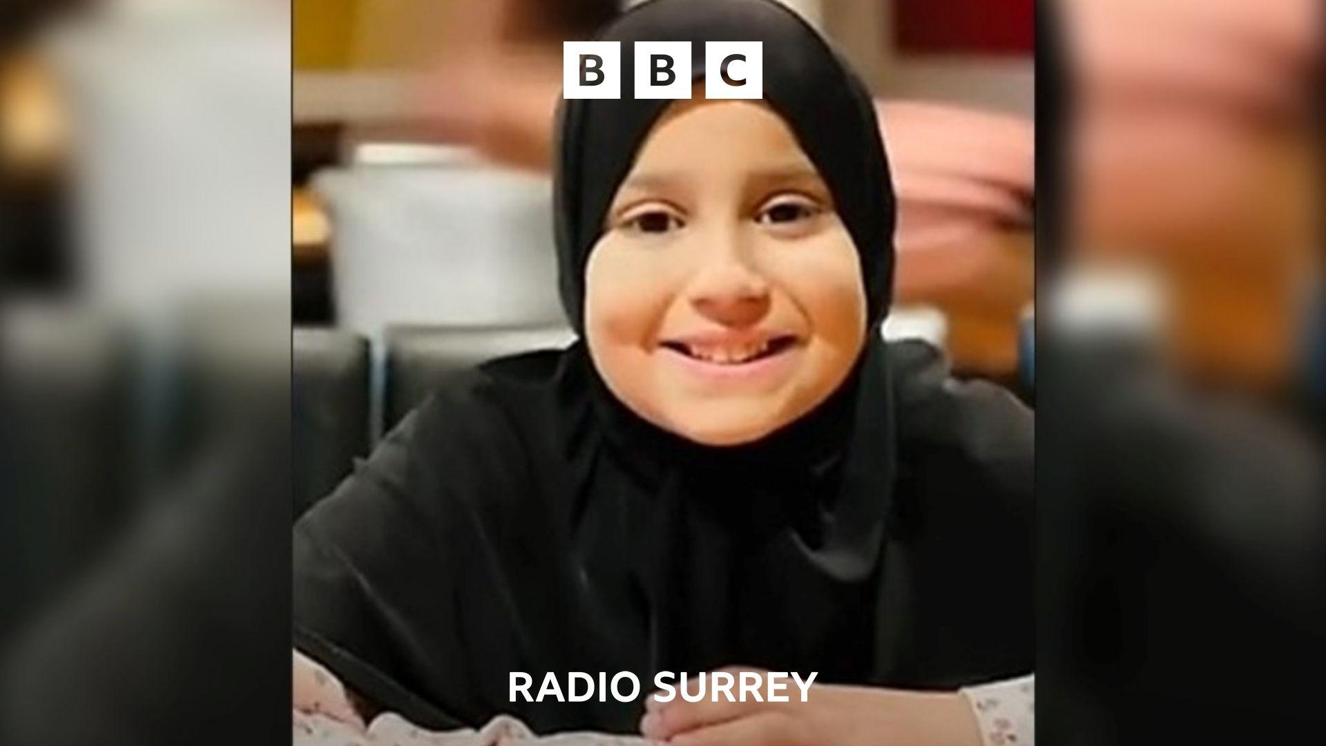 A girls smiles at the camera. She is wearing a hijab and her hands rest on the desk in front of her.