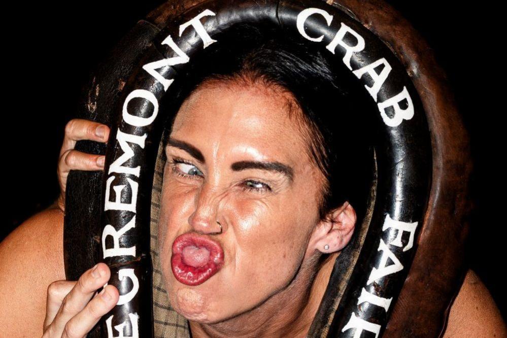 Claire Lister is distorting her face, going cross-eyed and sticking her tongue out. She has black hair and a nose ring. Her hands are placed on a large black horse collar put around her beck which says, in white lettering, Egremont Crab Fair.