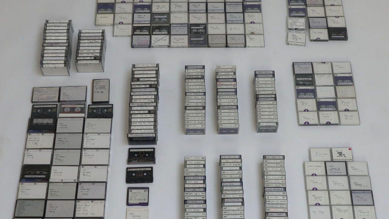 Dozens of tapes are grouped together and laid out on a white surface. There is small hand written notes on each tape.