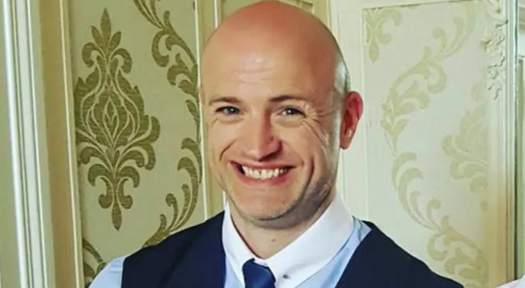Shows a man with bald head smiling. He is wearing a blue shirt with white collar, blue tie and blue waistcoat.