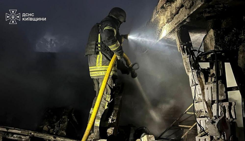 A firefighter wearing heavy duty black overalls with fluorescent green stripes holds a hosepipe next to a damaged buildings, which is at the scene of a Russian strike in the Kyiv region.