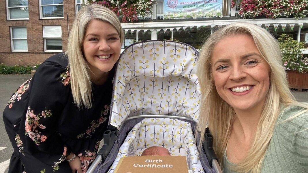 We spent £23,000 on fertility treatment – now our embryos will be destroyed In the middle of the photo is a pram. In the bassinet is a baby, you can't see his face but you can see the side of face. The outside of the pram is grey and the inside is white, there are grey trees with yellow balls coming off of them on the inside. On top of the bassinet is a brown envelope with 'Birth Certificate' written on it the top in white writing. There are two women, one sitting either side of the pram. The woman on the left is bending down next to the pram, wearing a black dress with pink and red flowers on it, her blonde hair is down and she is smiling. The woman on the right is crouching next to the pram, taking the photo. She is wearing a green top, her blonde hair is down and she smiling.