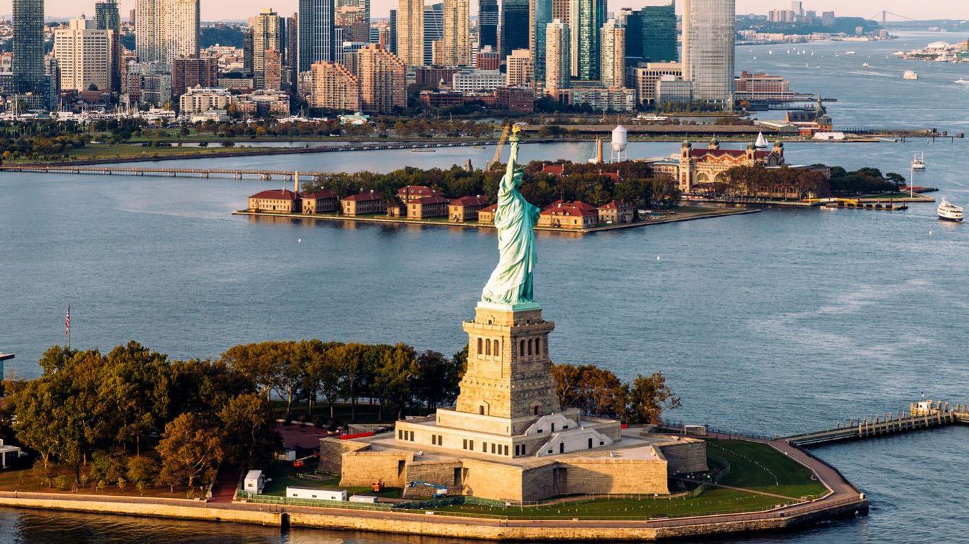 An aerial shot of Statue of Liberty and waters surrounding it with New York skyline in background