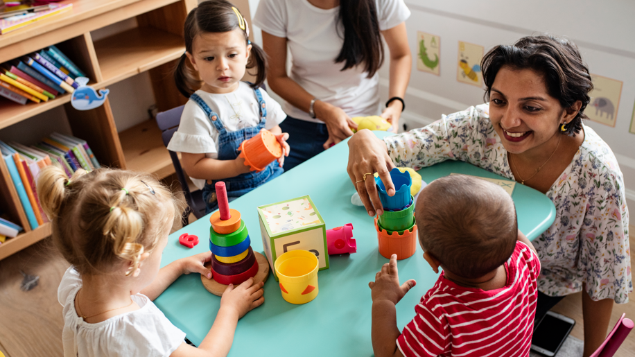 Children playing on a table with building toys. A woman plays with them and smiles.