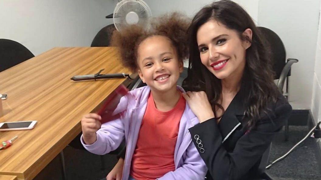 A little girl with bush hair, a purple jacket and red shirt, smiles as she sits on Cheryl's knee - Cheryl is also smiling and has long dark hair and is wearing a black jacket. They are sitting next to a desk with various items on it, in an office room.